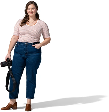 A woman in a tan pullover and blue jeans poses with her camera